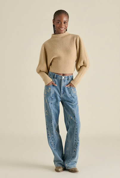 A model wearing a beige ribbed mock-neck cropped sweater and light-wash blue wide-leg jeans, standing against a solid light beige background.