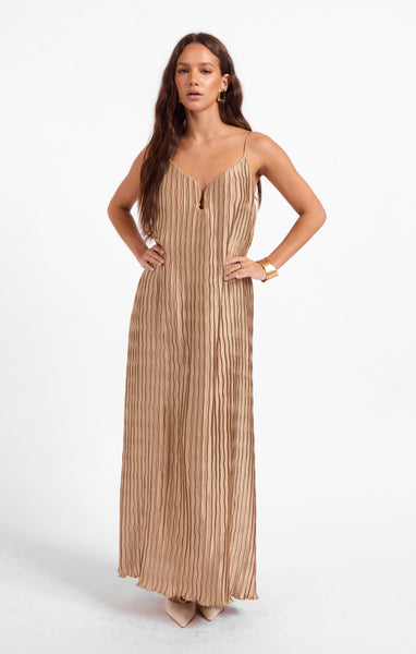 A woman modeling a sleeveless, floor-length pleated maxi dress in a warm beige/tan color, standing against a plain white studio background.