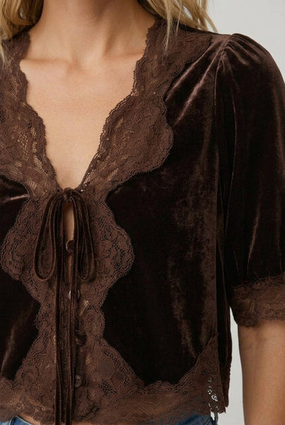 A woman wearing a dark brown velvet short-sleeve top with lace trim and a front tie detail, paired with blue jeans, against a light gray studio background.