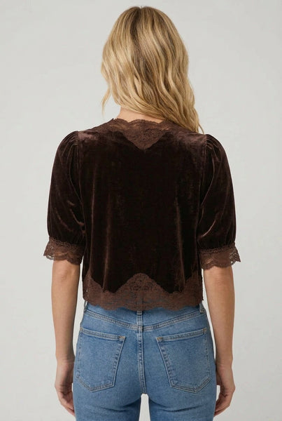 A woman wearing a dark brown velvet short-sleeve top with lace trim and a front tie detail, paired with blue jeans, against a light gray studio background.