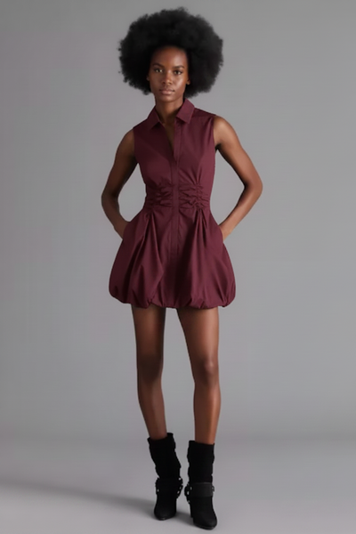 A model is wearing a black sleeveless mini dress with a buttoned collar and a gathered waist, paired with black ankle boots, against a solid gray studio background.