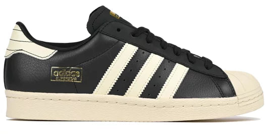 Adidas Superstar sneaker in black leather with cream-colored three stripes, a cream rubber toe cap, and a cream midsole, featuring a gold-toned metallic Adidas Originals logo on the side.
